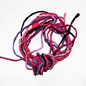 Può includere: Un groviglio di fili colorati nei toni del rosa, viola e nero su sfondo bianco. I fili sono di spessori e consistenze variabili, creando un'esposizione visivamente interessante. Ideale per progetti di artigianato.