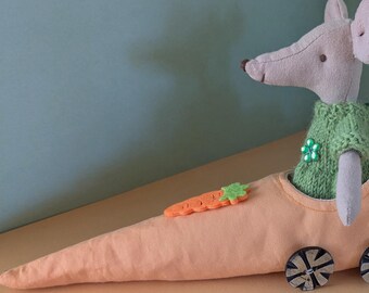 Könnte beinhalten: Eine graue Plüschmaus mit einem grünen Pullover sitzt in einem orangenen Plüsch-Karottenauto mit schwarzen Rädern. Das Auto hat einen grünen Stoffschwanz.
