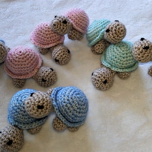 Könnte beinhalten: Sechs gehäkelte Schildkrötenspielzeuge in verschiedenen Farben, darunter rosa, blau und grün. Die Schildkröten haben braunes Garn für ihre Beine und Köpfe.