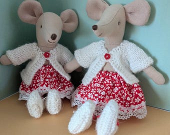 Könnte beinhalten: Zwei handgefertigte Mauspuppen. Jede Puppe hat einen beigefarbenen Kopf mit großen Ohren, einen weißen Strickcardigan und einen roten Rock mit Blumenmuster und Spitzenbesatz. Die Puppen haben weiße Strickfüße und einen roten Knopfverschluss am Cardigan.