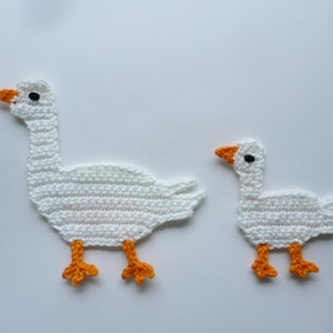 Könnte beinhalten: Zwei gehäkelte Gänse, eine größer als die andere, sind weiß mit orangefarbenen Schnäbeln und Füßen. Sie sind aus weißem Garn gefertigt und haben eine strukturierte Oberfläche.