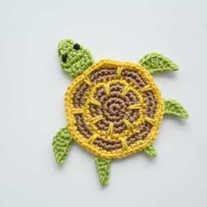 Könnte beinhalten: Ein gehäkelter Schildkröten-Untersetzer mit braunem und gelbem Zentrum und grünen gehäkelten Beinen und Kopf.