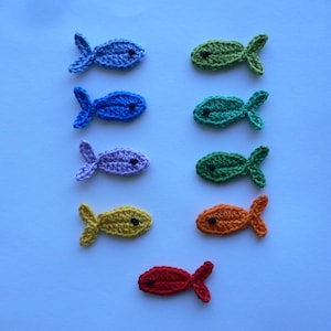 Könnte beinhalten: Sieben gehäkelte Fische in verschiedenen Farben, darunter Blau, Grün, Lila, Gelb, Orange und Rot. Die Fische sind vertikal auf einem hellblauen Hintergrund angeordnet.