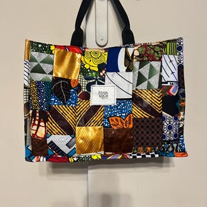 Peut inclure: Un sac fourre-tout patchwork avec une variété de carrés de tissu colorés. Le sac a des poignées noires et le texte "FAMA LOOK" sur une étiquette blanche. Le sac est accroché à une porte blanche.