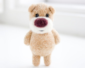Pode incluir: Um pequeno urso de peluche marrom de feltro com pelo branco ao redor do nariz e um nariz bordô. O urso está em pé sobre uma superfície branca.