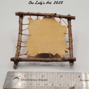 May include: A small, handcrafted wooden frame with a tan leather insert. The frame is constructed from twigs and tied together with twine. The text "One Lady's Art 2025" is visible. A stainless steel ruler is in the foreground.