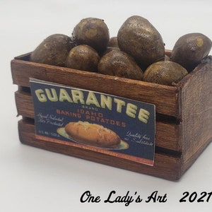 May include: A miniature wooden crate filled with brown potatoes. The crate has a label that reads "GUARANTEE BRAND IDAHO BAKING POTATOES." The crate is made of dark brown wood and sits on a white surface. The text "One Lady's Art 2021" is at the bottom.
