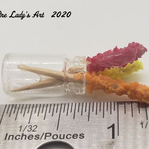 May include: A small glass jar with a clear lid containing two wooden sticks and three colorful crystal-like objects. The jar is sitting on a ruler marked in inches.
