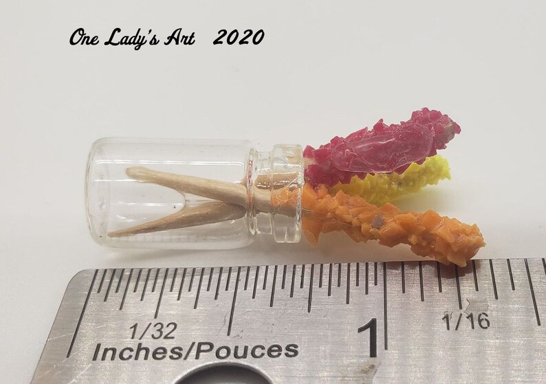May include: A small glass jar with a clear lid containing two wooden sticks and three colorful crystal-like objects. The jar is sitting on a ruler marked in inches.