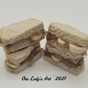 May include: A stack of four miniature peanut butter and banana sandwiches made of polymer clay. The sandwiches are brown and beige with white banana slices.