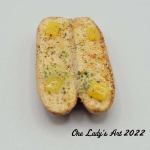 Pode incluir: Uma escultura em miniatura de argila polimérica, feita à mão, com tema de comida, que representa um pão de alho com manteiga derretida e ervas. O pão está partido ao meio e o interior está recheado com manteiga amarela e ervas verdes.