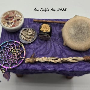 May include: A collection of spiritual items arranged on a small purple table. Includes a dreamcatcher, a small bowl, a shell, a smudge stick, a small drum, and a small item with text. The text "One Lady's Art 2025" is visible.
