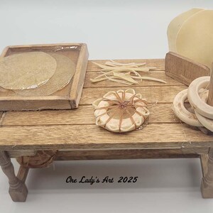 May include: A miniature wooden table displays various craft supplies. Items include a tray with two circular pieces, a bundle of thin sticks, a small round object tied with twine, and a stack of wooden rings. The table has a distressed finish.