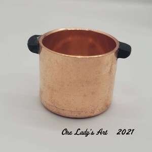 May include: A small copper pot with black plastic handles. The pot is about 2 inches tall and 2 inches wide.