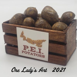 May include: A small wooden crate filled with brown potatoes. The crate has a label that says "P.E.I. POTATOES" and a small image of Prince Edward Island. The text "One Lady's Art 2021" is printed below the crate.