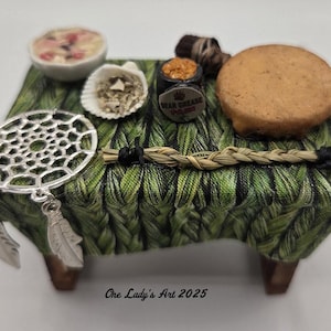 May include: Miniature table with a green woven fabric tablecloth. The table holds a dreamcatcher, a bowl of food, a seashell, a jar labeled "Bear Grease", a biscuit, and a bundle of twigs. The table is made of wood.
