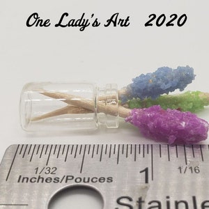 May include: Three colorful rock candy sticks in a small glass jar with a wooden skewer. The candy is blue, green, and purple. The jar is sitting on a ruler with the measurement 1/32 inches.