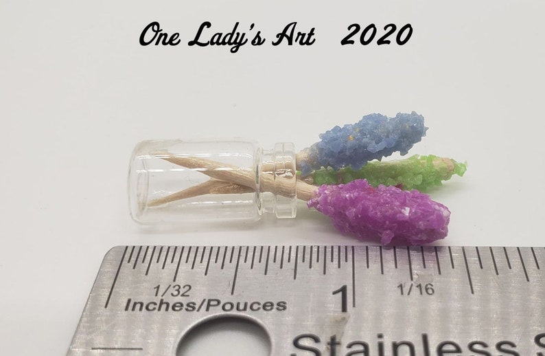 May include: Three colorful rock candy sticks in a small glass jar with a wooden skewer. The candy is blue, green, and purple. The jar is sitting on a ruler with the measurement 1/32 inches.