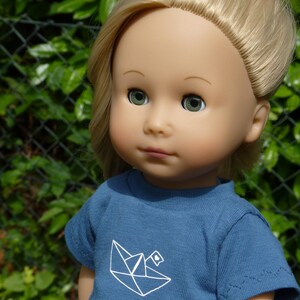May include: A doll with blonde hair and green eyes wearing a blue t-shirt with a white origami boat design. The doll is a light skin tone and is set against a green background.
