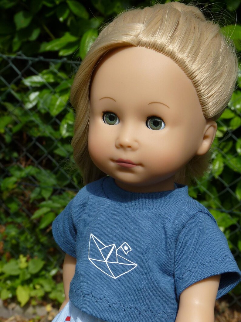 May include: A doll with blonde hair and green eyes wearing a blue t-shirt with a white origami boat design. The doll is a light skin tone and is set against a green background.