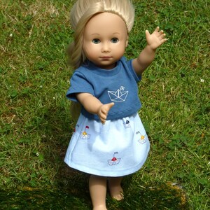 May include: A doll with blonde hair wearing a blue t-shirt with a white paper boat graphic and a light blue skirt decorated with sailboat designs. The doll is standing on green grass.