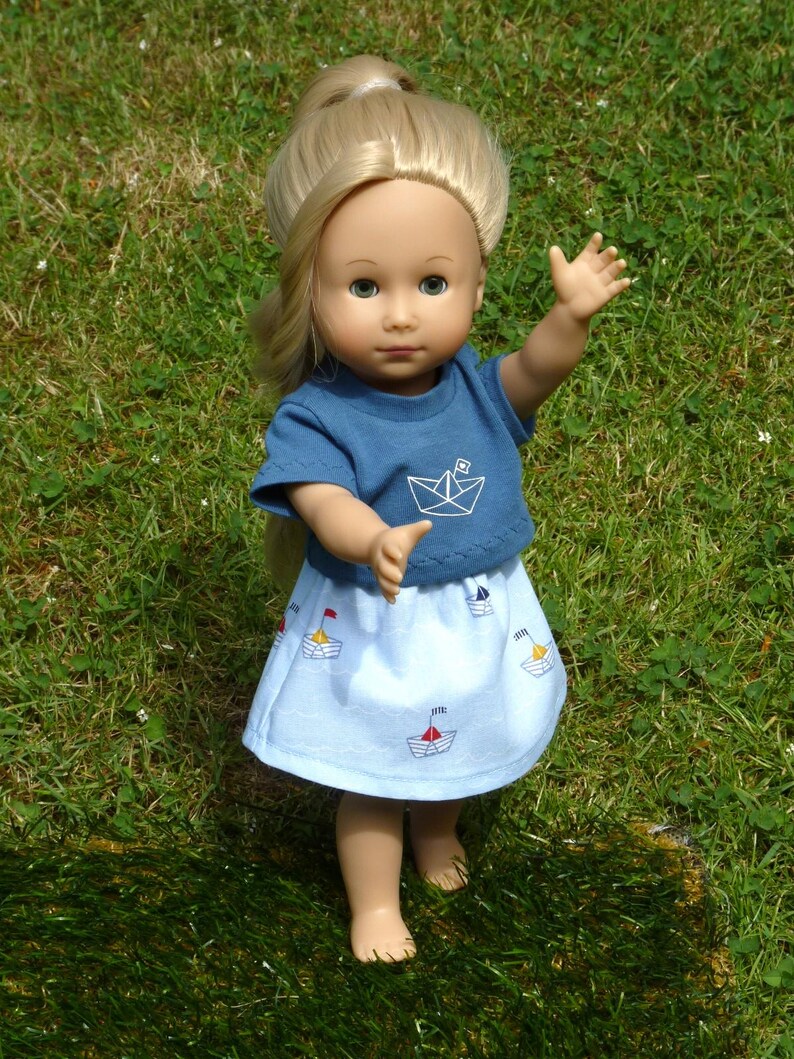 May include: A doll with blonde hair wearing a blue t-shirt with a white paper boat graphic and a light blue skirt decorated with sailboat designs. The doll is standing on green grass.