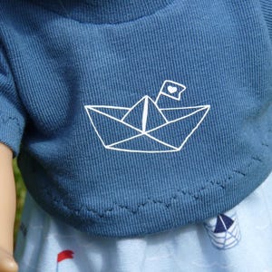 May include: A blue t-shirt with a white paper boat graphic and a heart flag. The shirt is part of a doll outfit, paired with a light blue skirt featuring boat and flag patterns. The outfit is designed for a doll.