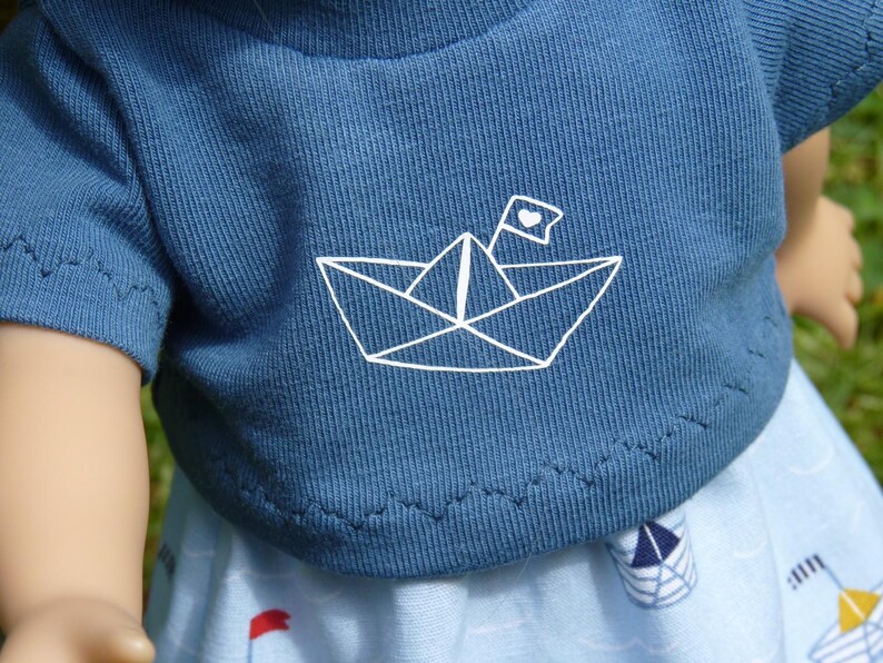 May include: A blue t-shirt with a white paper boat graphic and a heart flag. The shirt is part of a doll outfit, paired with a light blue skirt featuring boat and flag patterns. The outfit is designed for a doll.