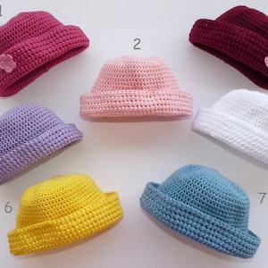 Può includere: Sette cappelli a secchiello all'uncinetto in vari colori: fucsia, rosa chiaro, bordeaux, lavanda, bianco, giallo e blu. Alcuni cappelli hanno un piccolo ornamento floreale. I cappelli sono esposti su sfondo bianco.