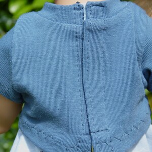 May include: Close-up of a blue short-sleeved shirt with a zipper down the back. The shirt is made of a textured fabric and has a high neckline. The sleeves are hemmed with a zig-zag stitch.