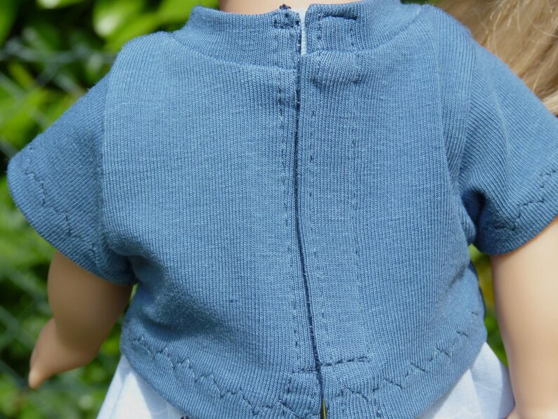 May include: Close-up of a blue short-sleeved shirt with a zipper down the back. The shirt is made of a textured fabric and has a high neckline. The sleeves are hemmed with a zig-zag stitch.