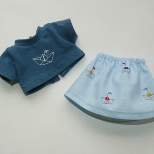 May include: A blue doll outfit featuring a short-sleeved top with a white paper boat design and a light blue skirt with embroidered sailboats. The skirt has a wavy pattern, and the outfit is made of soft fabric.