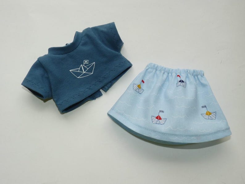 May include: A blue doll outfit featuring a short-sleeved top with a white paper boat design and a light blue skirt with embroidered sailboats. The skirt has a wavy pattern, and the outfit is made of soft fabric.