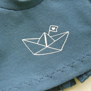 May include: A close-up of a teal-colored t-shirt featuring a white paper boat design with a heart on the flag. The shirt has a ribbed texture and a raw hemline. The design is simple and minimalist.