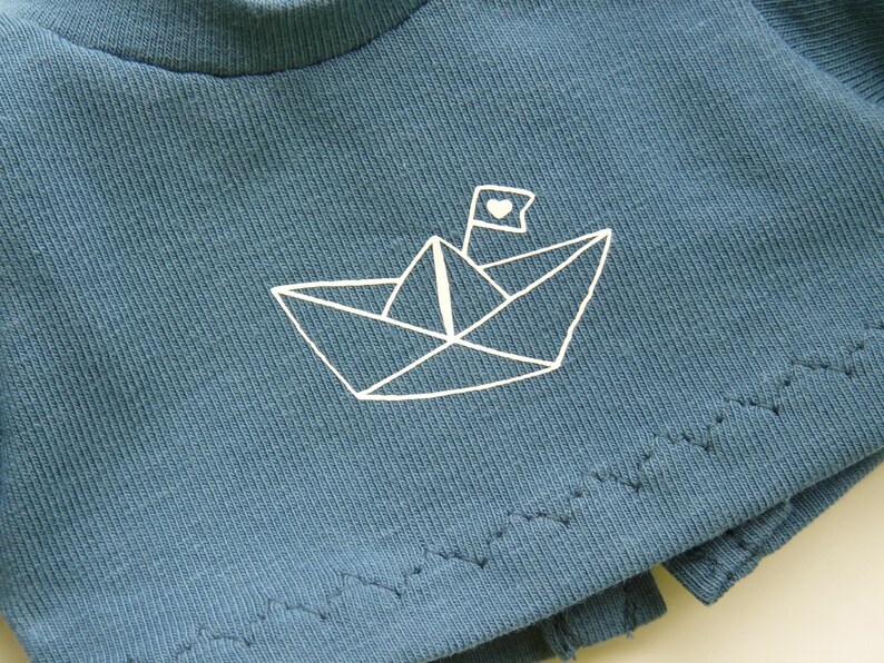 May include: A close-up of a teal-colored t-shirt featuring a white paper boat design with a heart on the flag. The shirt has a ribbed texture and a raw hemline. The design is simple and minimalist.