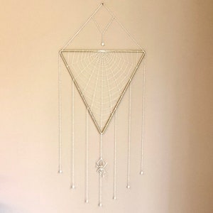 Könnte beinhalten: Ein Traumfänger in Form eines weißen Spinnennetzes mit einem goldenen dreieckigen Rahmen und weißen Perlenketten, die von unten hängen. Der Traumfänger hängt an einer beigefarbenen Wand.