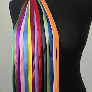 Pode incluir: Uma coleção de fitas de cetim verticais num espectro de cores que inclui laranja, rosa claro, azul, amarelo, bordeaux, roxo, preto, vermelho, verde e castanho, drapeadas sobre um torso de manequim preto para mostrar a textura lisa e brilhante.