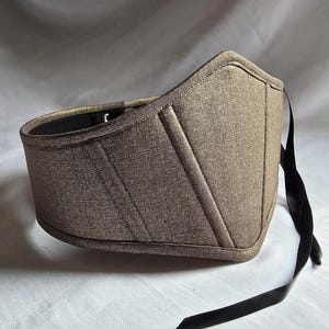 May include: A metallic bronze-colored corset with a structured design. The corset features a curved shape, diagonal seams, and a black ribbon tie. The interior is black, and the overall aesthetic is elegant and refined.