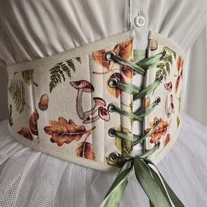 Peut inclure: Corset crème avec un motif automnal de champignons, de feuilles et de glands. Il présente un laçage vert à l'avant et une bordure crème. Le motif incorpore des nuances de marron, d'orange et de vert.