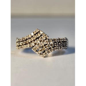 May include: A silver-toned ring featuring a bypass design with two overlapping bands. The surface of the ring is densely encrusted with numerous small, clear, round-cut stones that create a continuous sparkling texture across the entire visible front.