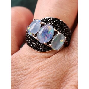 Könnte beinhalten: Ein silberner Ring mit drei ovalen Opalen. Die Opale sind hellblau mit schillernden Reflexen. Das Ringband ist mit kleinen schwarzen Steinen besetzt, was einen auffälligen Kontrast erzeugt. Der Ring wird an einem Finger getragen.