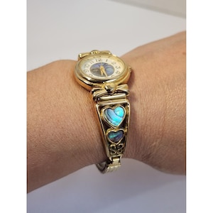 Op de afbeelding: Goudkleurig polshorloge met een ronde wijzerplaat en een band met hartvormige inlays met iriserende blauwe en groene patronen. De wijzerplaat toont de nummers 1 tot en met 12. Het horloge zit om een pols.