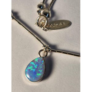 Op de afbeelding: Een zilveren ketting met een druppelvormige opaal hanger met iriserende blauwe en groene flitsen. De ketting bestaat uit dunne zilveren staafjes en kleine kraaltjes. Een ovaal metalen label aan de sluiting is gestempeld met 925 S.