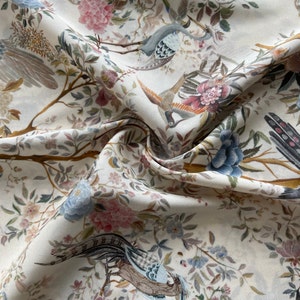 Può includere: Tessuto bianco sporco con un motivo floreale e di uccelli dettagliato. Il design presenta uccelli in volo e appollaiati sui rami, circondati da fiori rosa, blu e malva. Il tessuto ha un aspetto morbido e fluido.