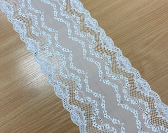 Op de afbeelding: Witte kanten afwerking met een geschulpte rand en een bloemenmotief. Het kant ligt diagonaal over een lichtbruin houten oppervlak. Het kant heeft een zigzagpatroon met kleine bloemendetails.
