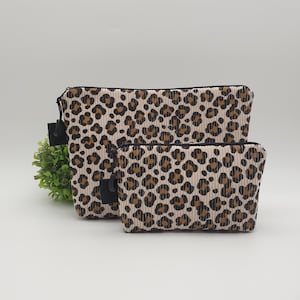 Könnte beinhalten: Zwei Kosmetiktaschen aus Cord mit Leopardenmuster. Die Taschen sind beige mit braunen Flecken und schwarzen Reißverschlüssen. Eine Tasche ist größer als die andere.