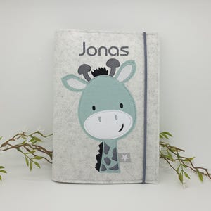 Könnte beinhalten: Eine hellgraue Filzhülle mit einem türkisgrünen Giraffen-Design und dem Namen "Jonas" auf der Vorderseite. Die Giraffe hat ein weißes Gesicht, schwarze Flecken und einen kleinen Stern. Ein graues Gummiband sichert die Hülle.