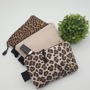 Könnte beinhalten: Drei rechteckige Kord-Etuis mit Reißverschluss. Zwei Etuis haben ein braun-schwarzes Leopardenmuster, während das mittlere Etui einfarbig beige ist. Jedes Etui verfügt über einen schwarzen Reißverschlussanhänger und einen kleinen goldenen Buchstabenanhänger.