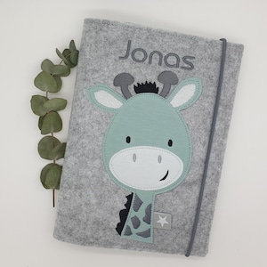 Könnte beinhalten: Graues Filz-Babybuch mit einer grünen und weißen Giraffen-Applikation und dem Namen Jonas, der auf der Vorderseite gestickt ist.