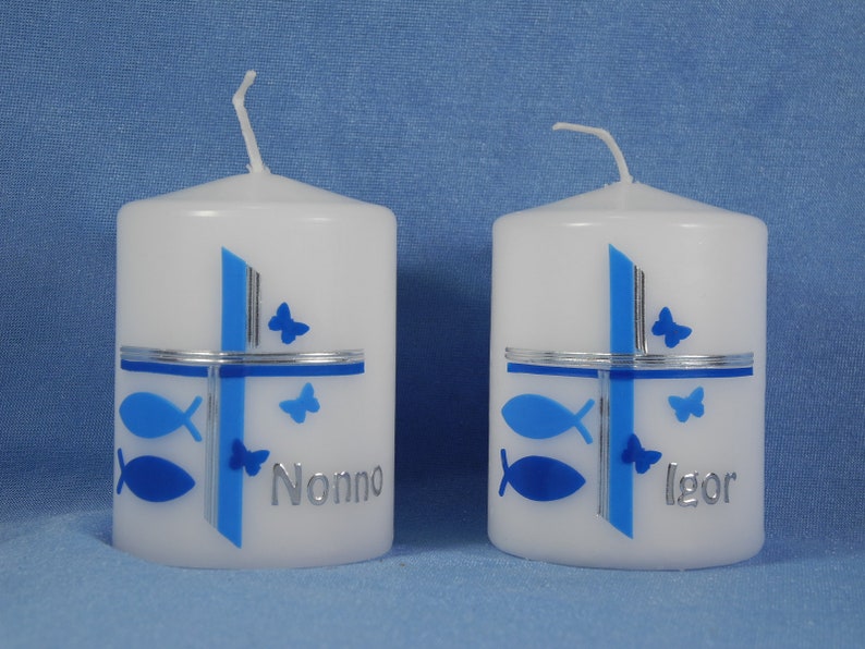 K&ouml;nnte beinhalten: Zwei wei&szlig;e Kerzen mit blauen und silbernen Designs. Die Kerzen haben ein Kreuz mit Fischen und Schmetterlingen. Eine Kerze sagt "Nonno" und die andere sagt "Igor".